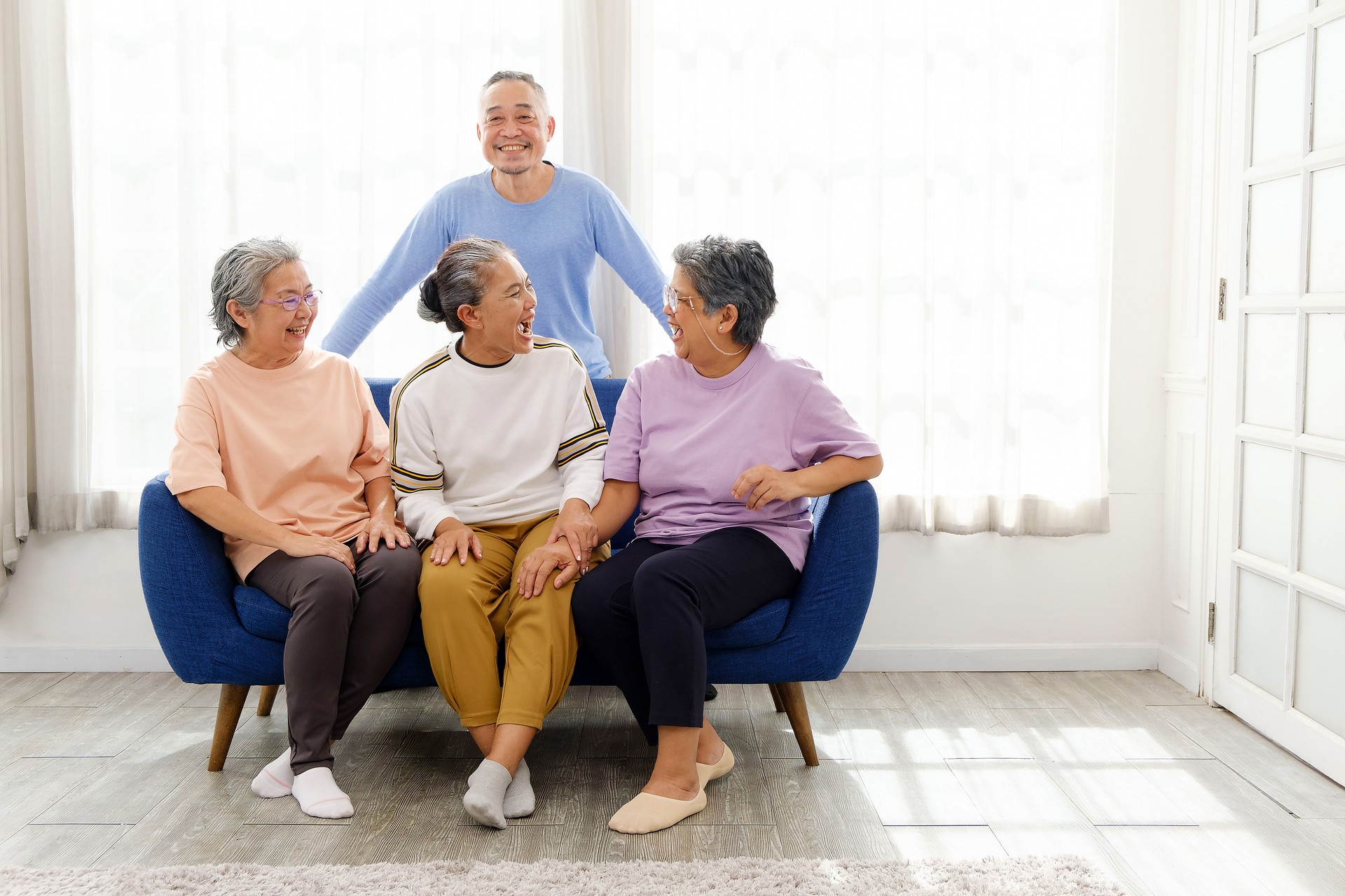 Four happy Asian senior friends enjoying laughter and bonding in a cozy home setting. Elderly man and three women sitting on a couch, embracing companionship, joy, and a healthy social lifestyle.